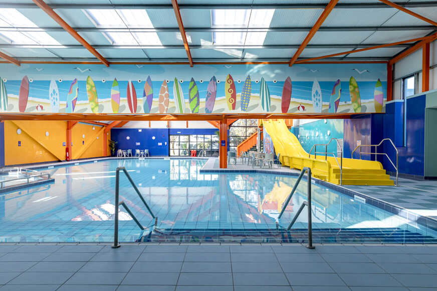Indoor swimming pool with a yellow slide and surfboard mural.