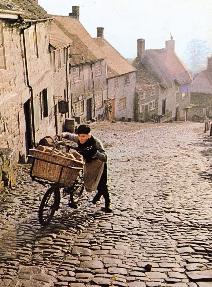 Boy pushing bread delivery bicycle up cobblestone hill.