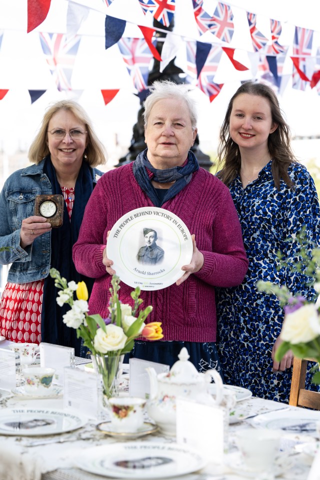 Three women at a table displaying a commemorative plate honoring Arnold Sharrocks, a WWII hero.