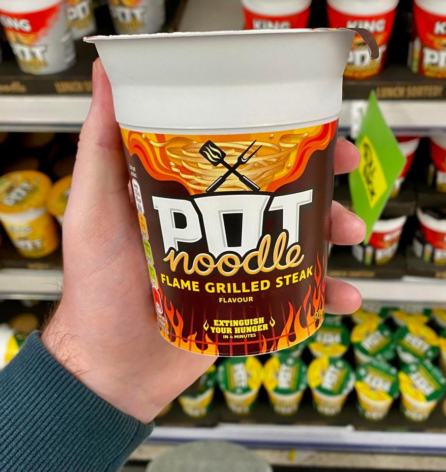 Hand holding a Pot Noodle cup, flame-grilled steak flavor.