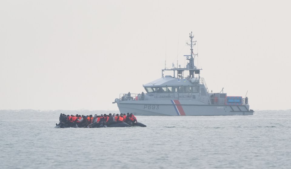 Migrants in a small boat being monitored by a larger vessel.