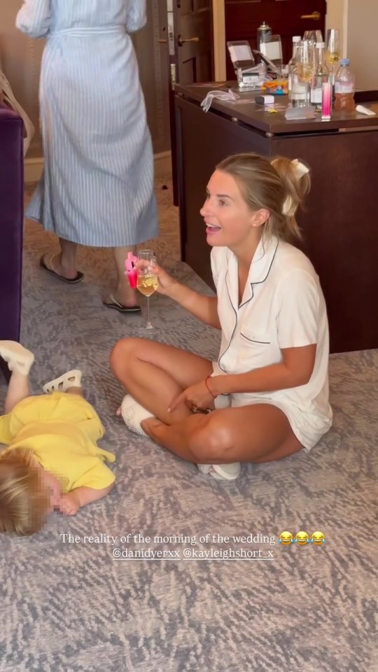 Woman in pajamas sits on the floor holding a glass of champagne while a baby lies on the floor nearby.