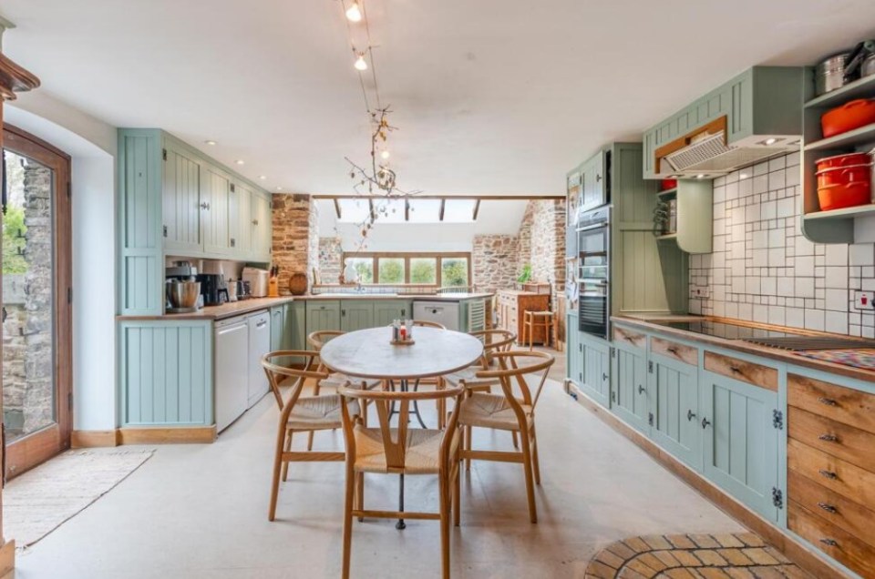 Kitchen with light green cabinets, wooden table and chairs, and exposed stone wall.