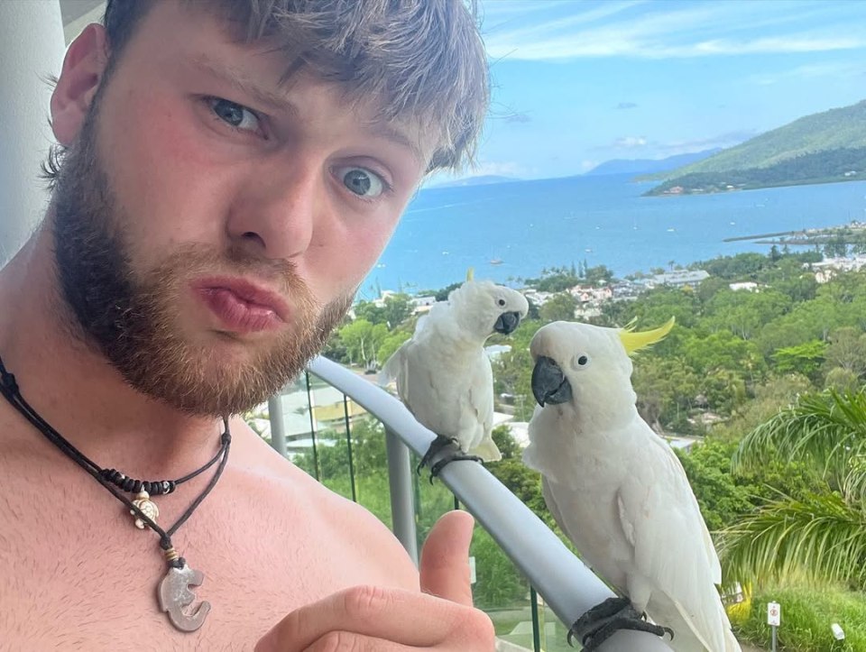 Selfie of a man with two white cockatoos on a balcony overlooking the ocean.