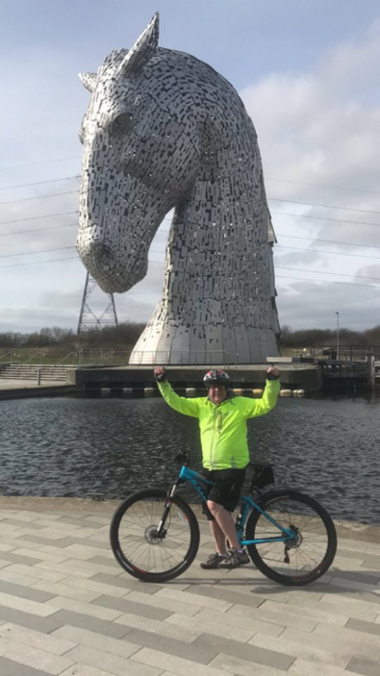 Cyclist posing with arms raised near a large horse head sculpture.