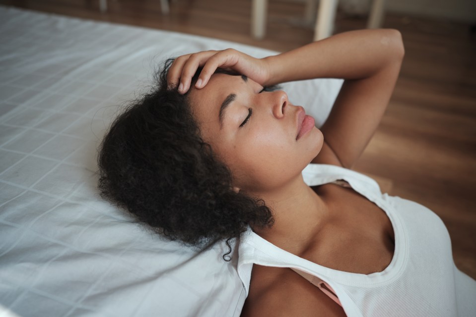 A young woman lies on her side with her eyes closed and hand on her forehead, appearing stressed or in pain.