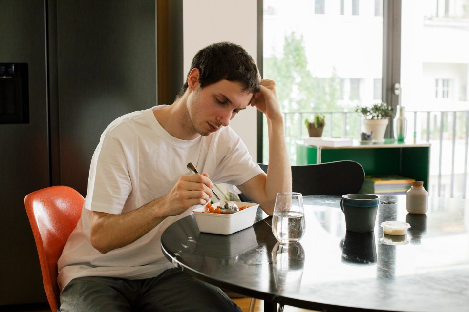 A young man sits at a table, looking sad while eating a meal.