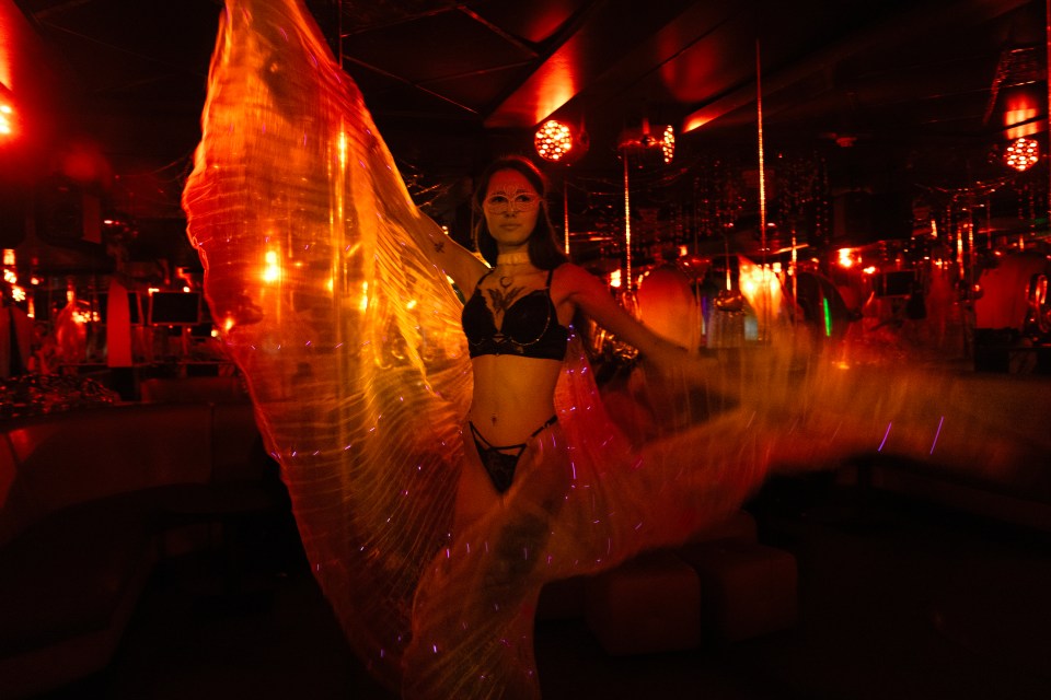 A dancer in a dimly lit nightclub in Kharkiv, Ukraine, wearing a mask and lingerie, poses with a glowing fabric.