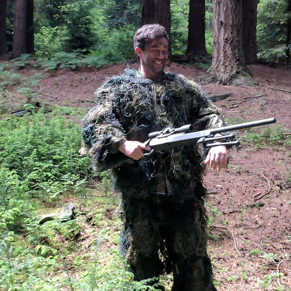 Actor in ghillie suit holding a rifle in a forest.