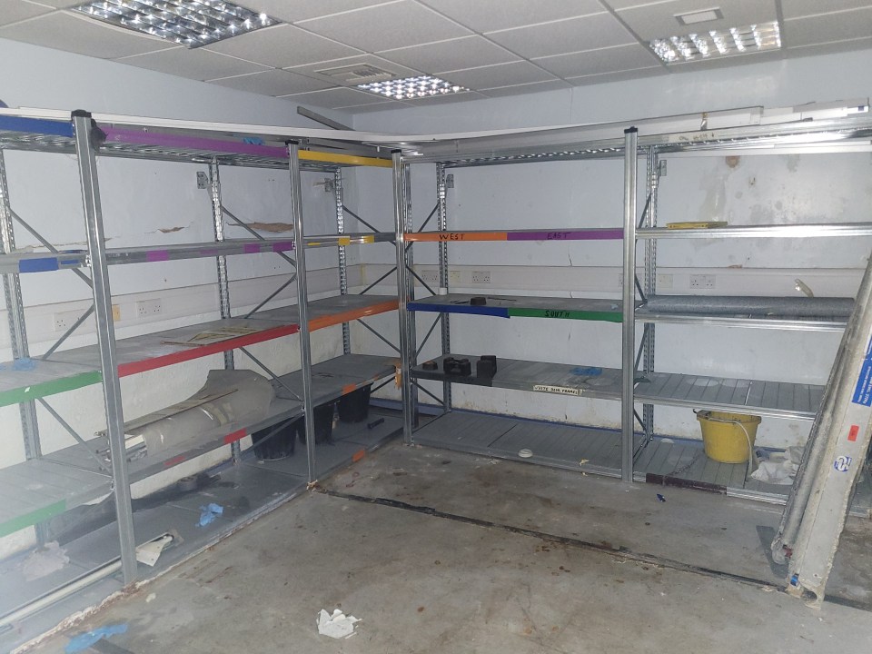 A cluttered storage room in a dilapidated hospital, with metal shelving units labeled "West," "East," and "South."