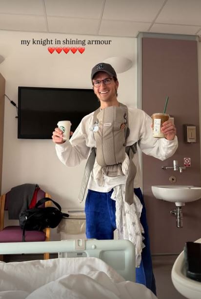 Man in hospital room holding baby carrier and coffees.