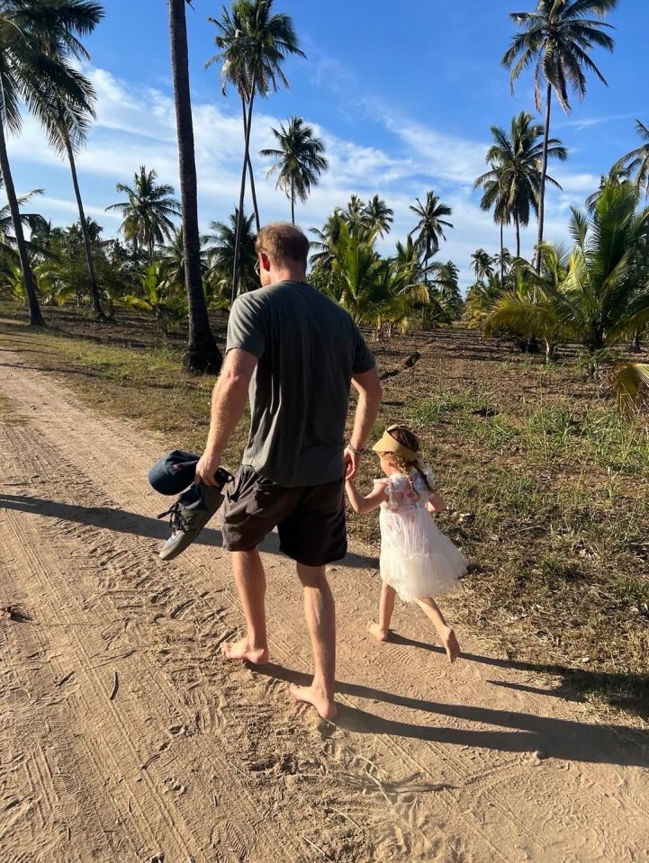 Prince Harry and Lilibet walking barefoot on a dirt road lined with palm trees.