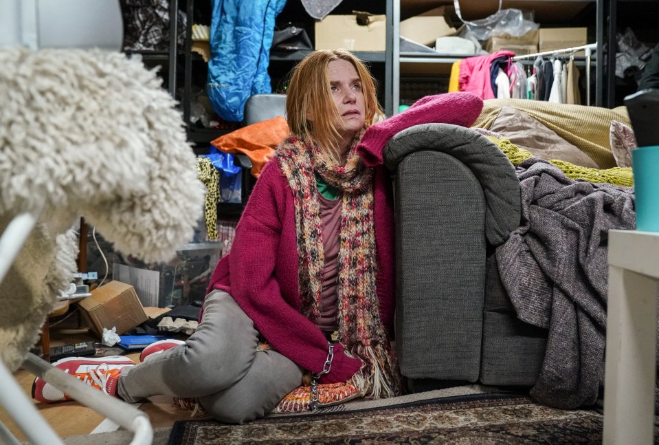 A woman sits on the floor in a cluttered room, chained to a sofa.