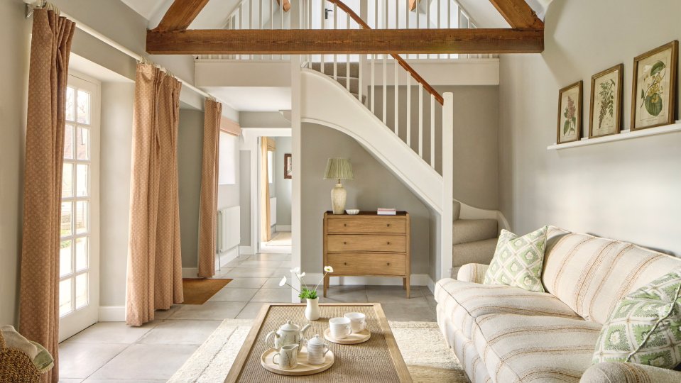 Interior view of a Cotswolds home's living room, featuring a curved staircase, a beige sofa, and a wooden chest of drawers.