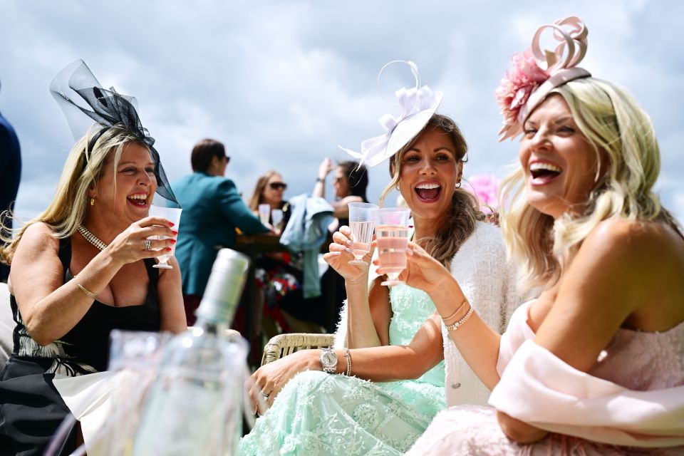 Three women in elegant hats at a horse race, toasting with drinks.