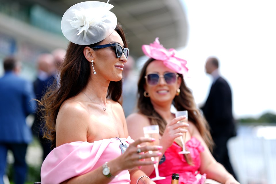 Two women in pink dresses and sunglasses at a horse race, drinking champagne.