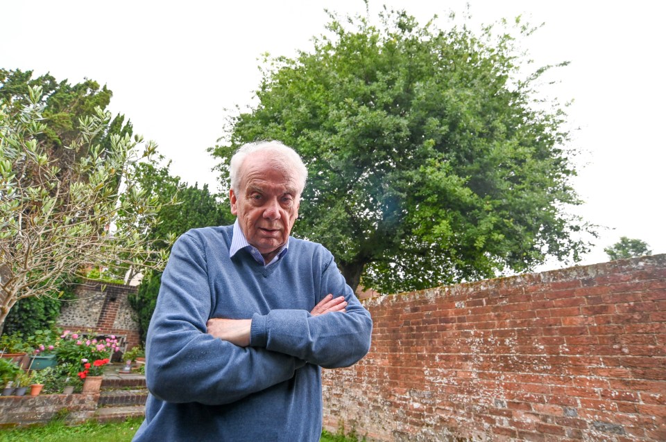 A man stands in his garden with arms crossed, looking at the camera, with a large tree behind him.