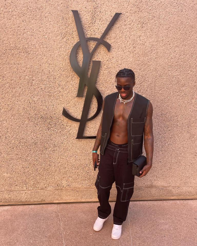 Man in black vest and pants standing in front of Yves Saint Laurent logo.