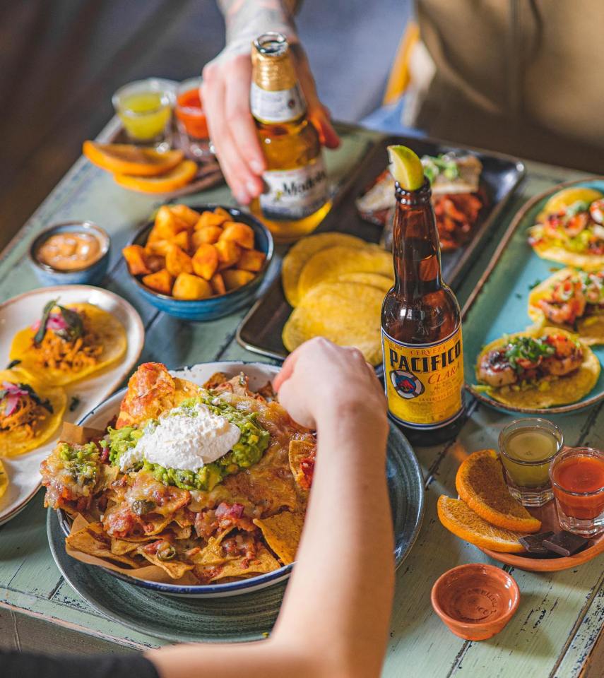 A table laden with Mexican food, including nachos, tacos, and potatoes, with bottles of beer.