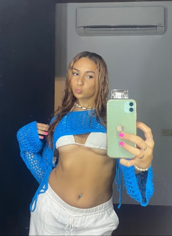 Selfie of a young woman wearing a blue crochet top and white pants.