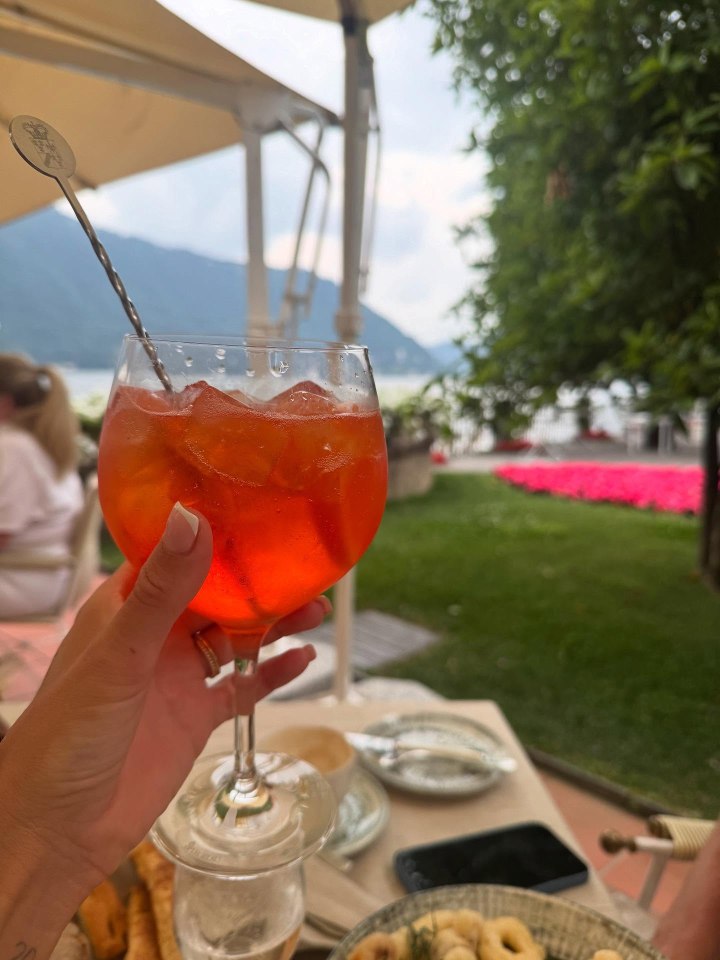 A hand holding an Aperol Spritz overlooking a lake.