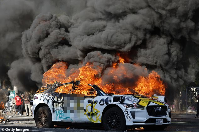 One protester repeatedly smashed the driver side window of a Waymo which had been surrounded by demonstrators as flames engulfed another car nearby