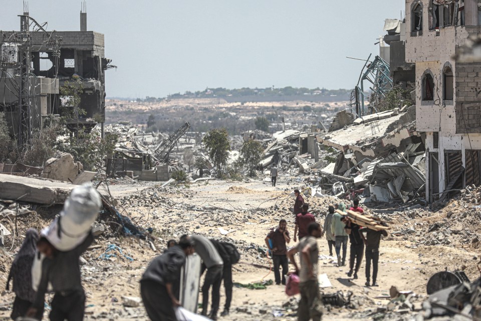 People carrying belongings walk through rubble-strewn streets amidst destroyed buildings.