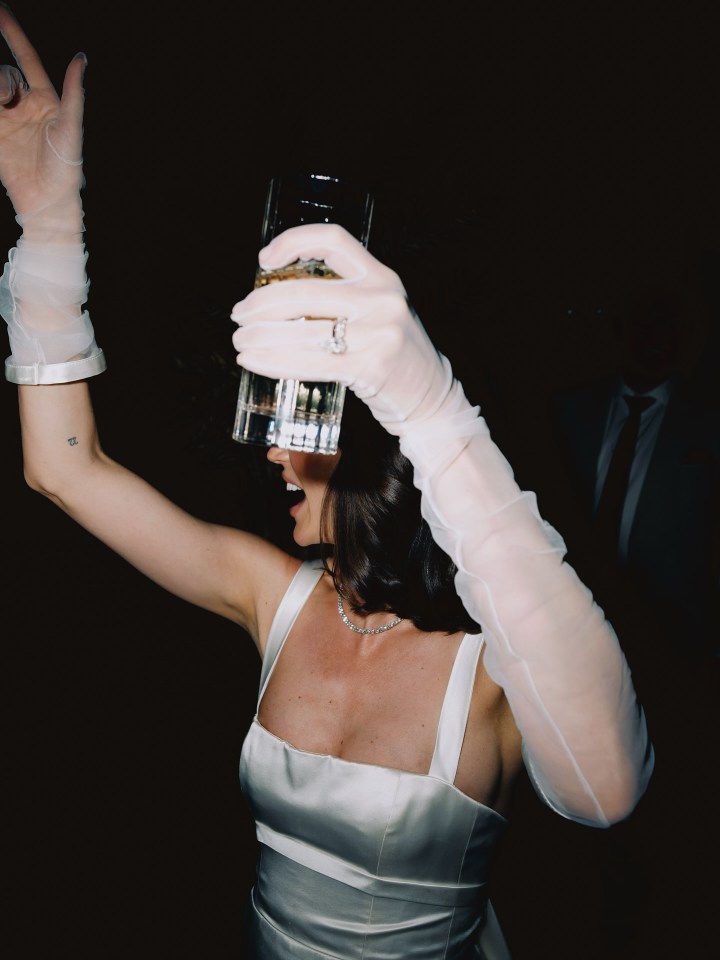 Bride in white dress holding a glass up to her face at a wedding reception.