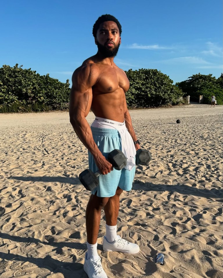 Muscular man lifting dumbbells on a beach.
