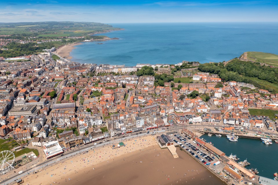 Aerial view of Scarborough, UK, showing the beach, town, and harbor on a sunny day.