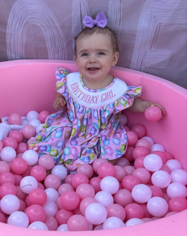Baby girl in a pink balloon-print dress sitting in a pink ball pit.