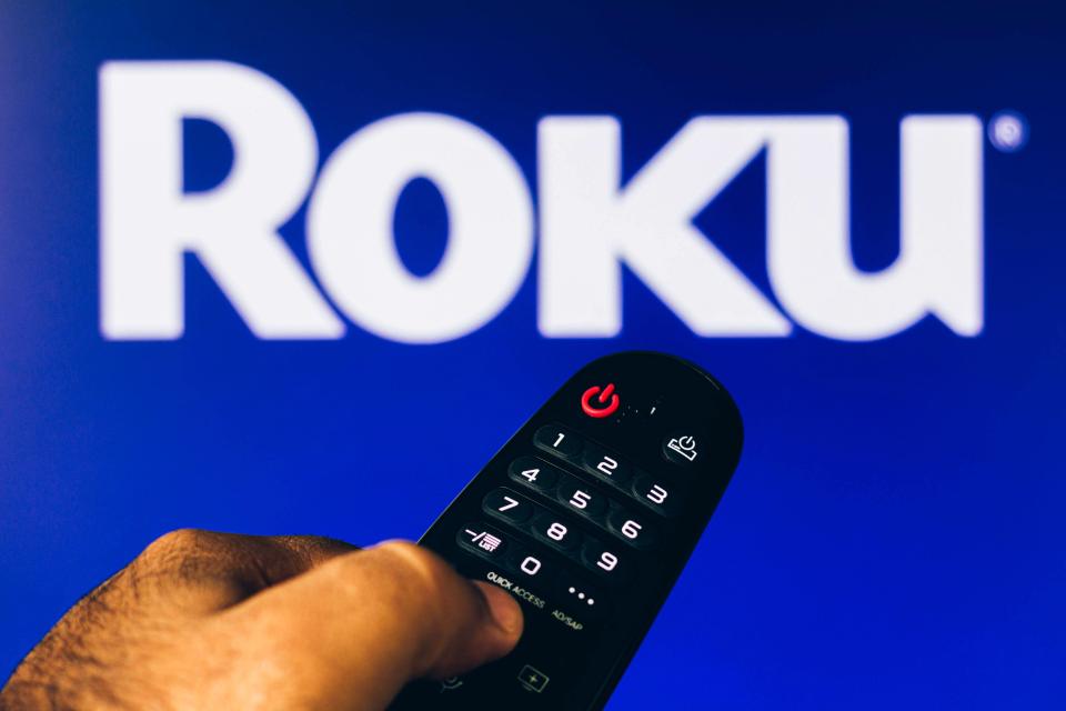Hand holding Roku remote control in front of Roku logo.