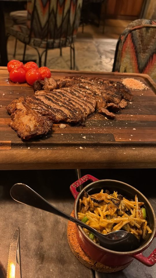 Sliced grilled steak on a wooden board with roasted tomatoes and a small dish of pasta.