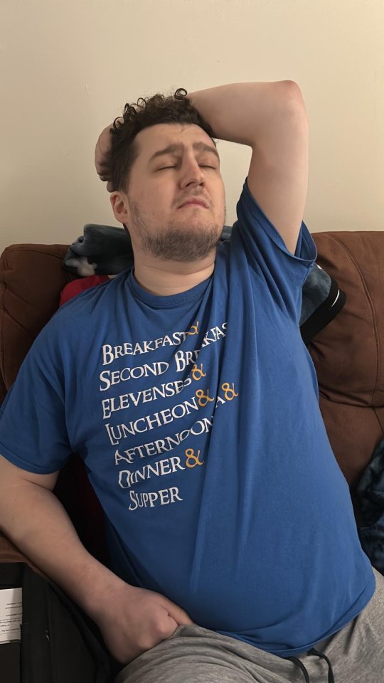 Man in blue shirt with meal times printed on it, sitting on a couch.