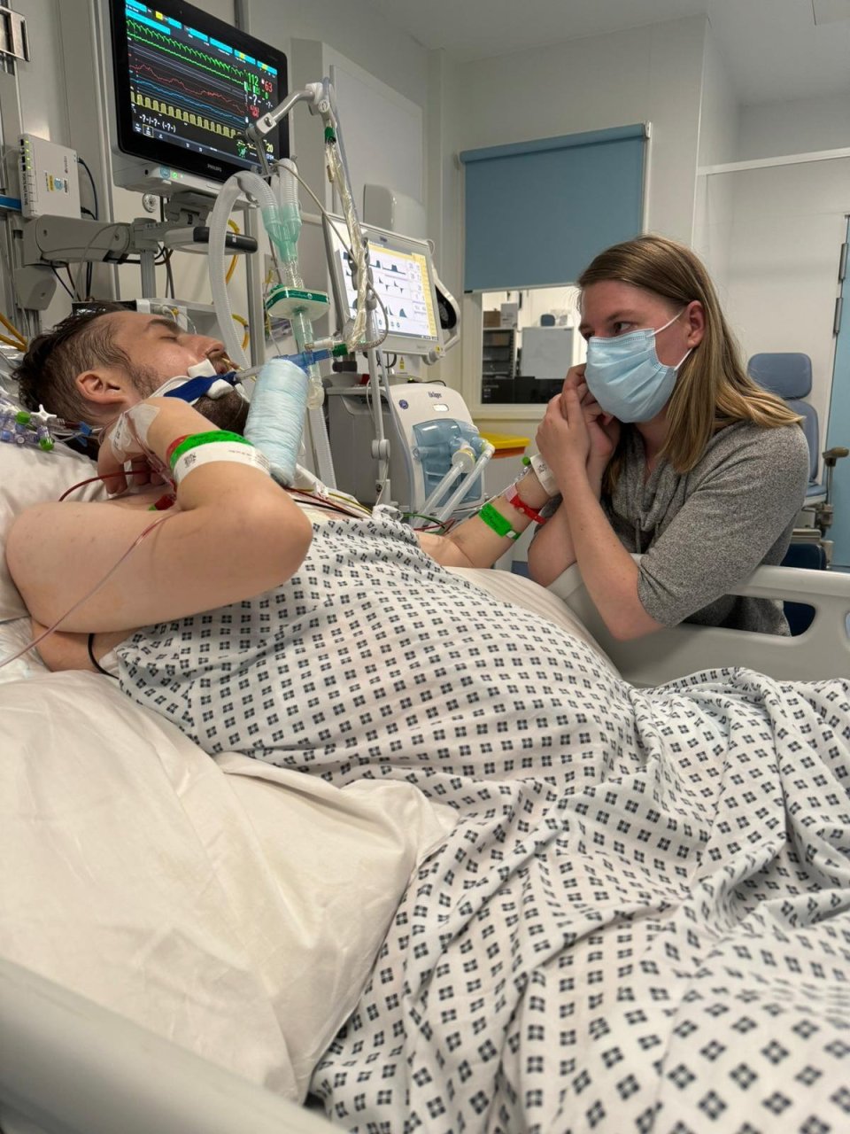 A woman holds her husband's hand while he lies in a hospital bed attached to medical equipment.