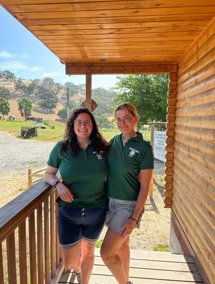 Two women in green Camp America staff shirts stand on a porch.