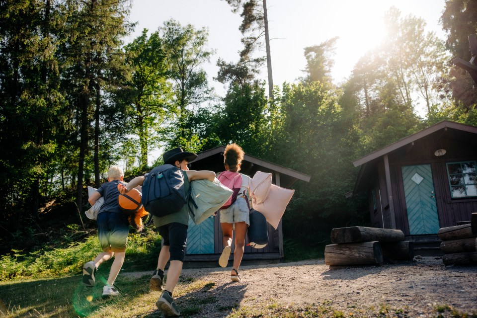 Three children carrying backpacks and pillows run toward a cabin in the woods.