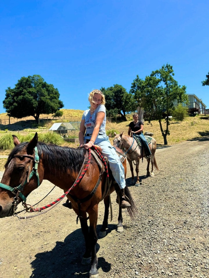 Two young women horseback riding on a gravel path.