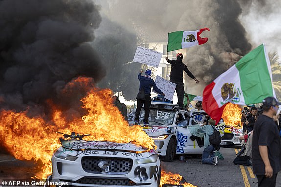 Demonstrators wave flags from atop a wrecked car, standing beside burning Waymo vehicles, as protesters clash with law enforcement in the streets surrounding the federal building during a protest following federal immigration operations in Los Angeles, California, on June 8, 2025. Demonstrators torched cars and scuffled with security forces in Los Angeles on June 8, as police kept protestors away from the National Guard troops President Donald Trump sent to the streets of the second biggest US city. (Photo by RINGO CHIU / AFP) (Photo by RINGO CHIU/AFP via Getty Images)