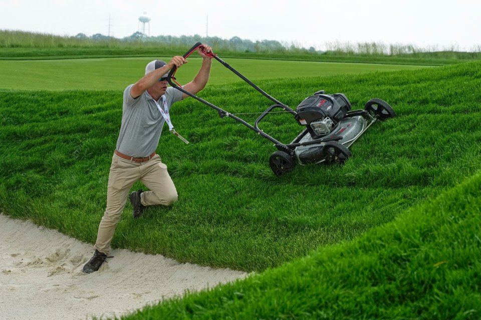 A groundskeeper struggles to pull a lawnmower up a steep grassy hill onto a sand trap.