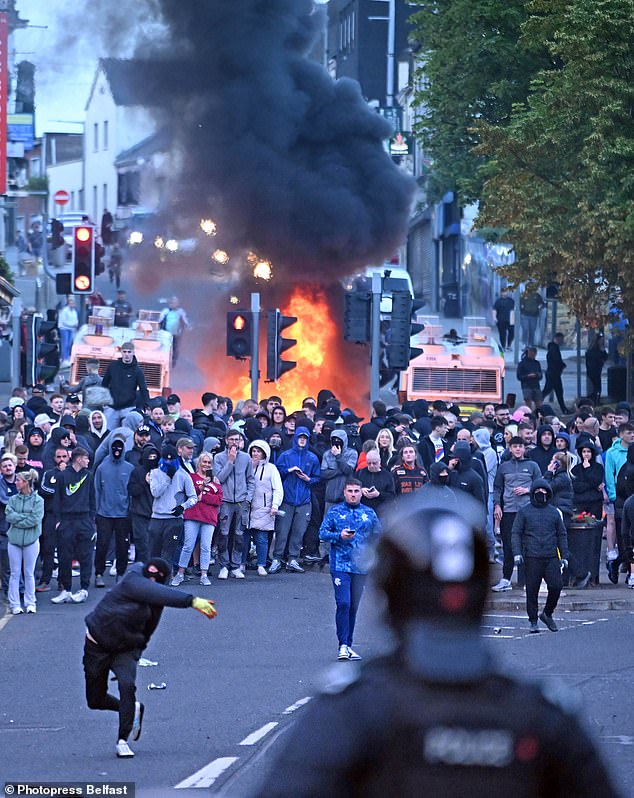 A thug wearing a balaclava hurls something at lines of police officers as cops came under attack for a second night