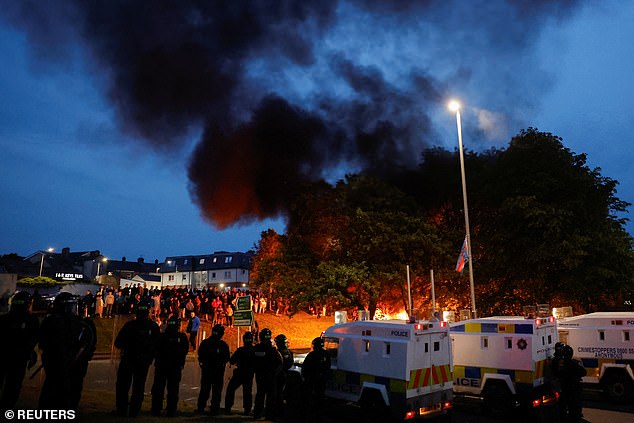 Police officers stand guard during the second night of riot as smoke belches into the sky