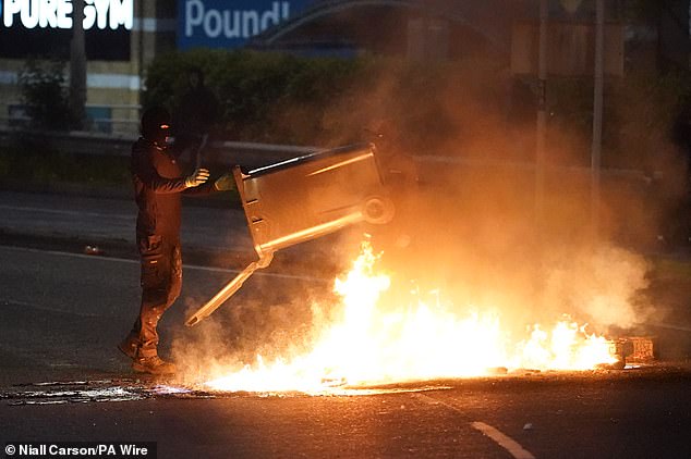 A masked thug throws a wheelie bin into the fire during the second evening of riots in the town