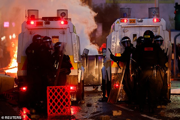Officers stand guard behind police vehicles as flames rise during the second evening of riots