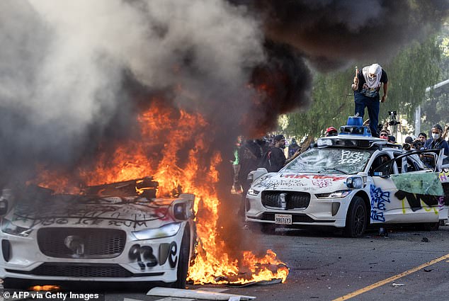 Demonstrators smash the windshield of a vehicle next to a burning Waymo vehicle as protesters clash with law enforcement in the streets surrounding the federal building during a protest following federal immigration operations in Los Angeles, California