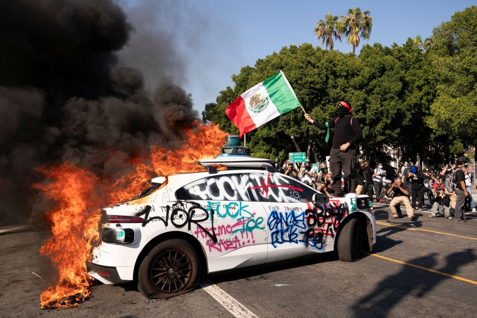 A burning car, vandalized with graffiti, at an immigration protest in Los Angeles. A person holds a Mexican flag.