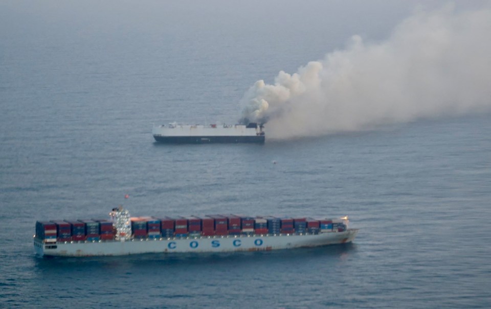 Aerial view of a cargo ship on fire at sea, with smoke billowing from it, and another cargo ship nearby.