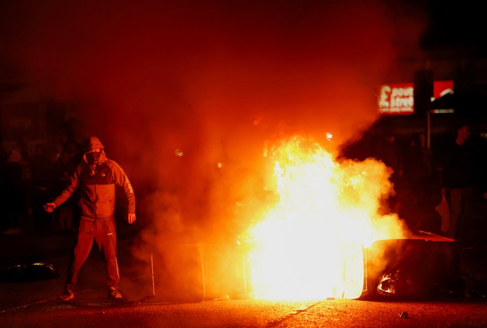 Masked demonstrator standing near a burning vehicle during riots.
