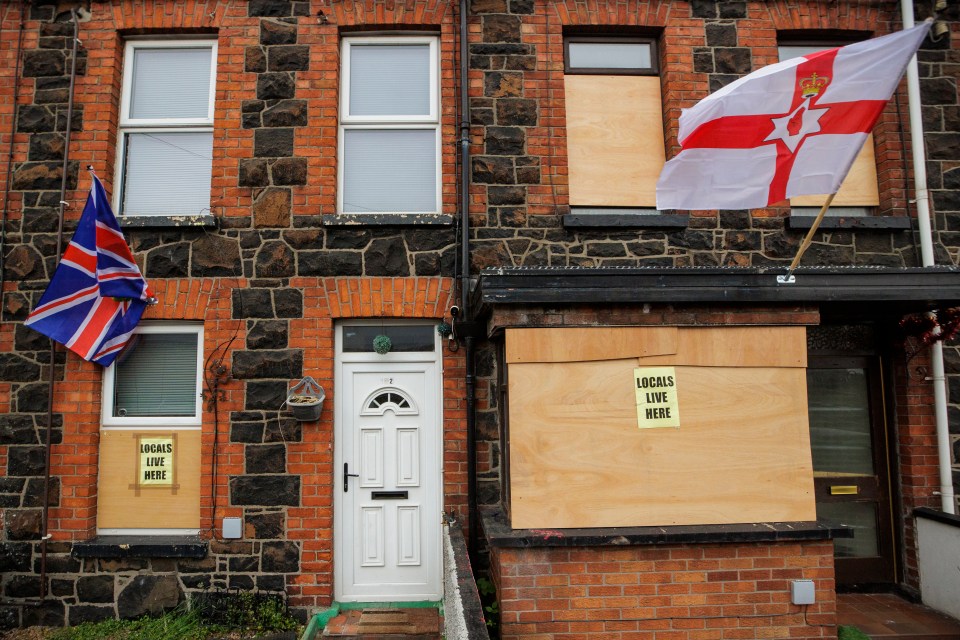 Ballymena houses with boarded windows and signs reading "Locals Live Here," along with UK and Ulster flags.