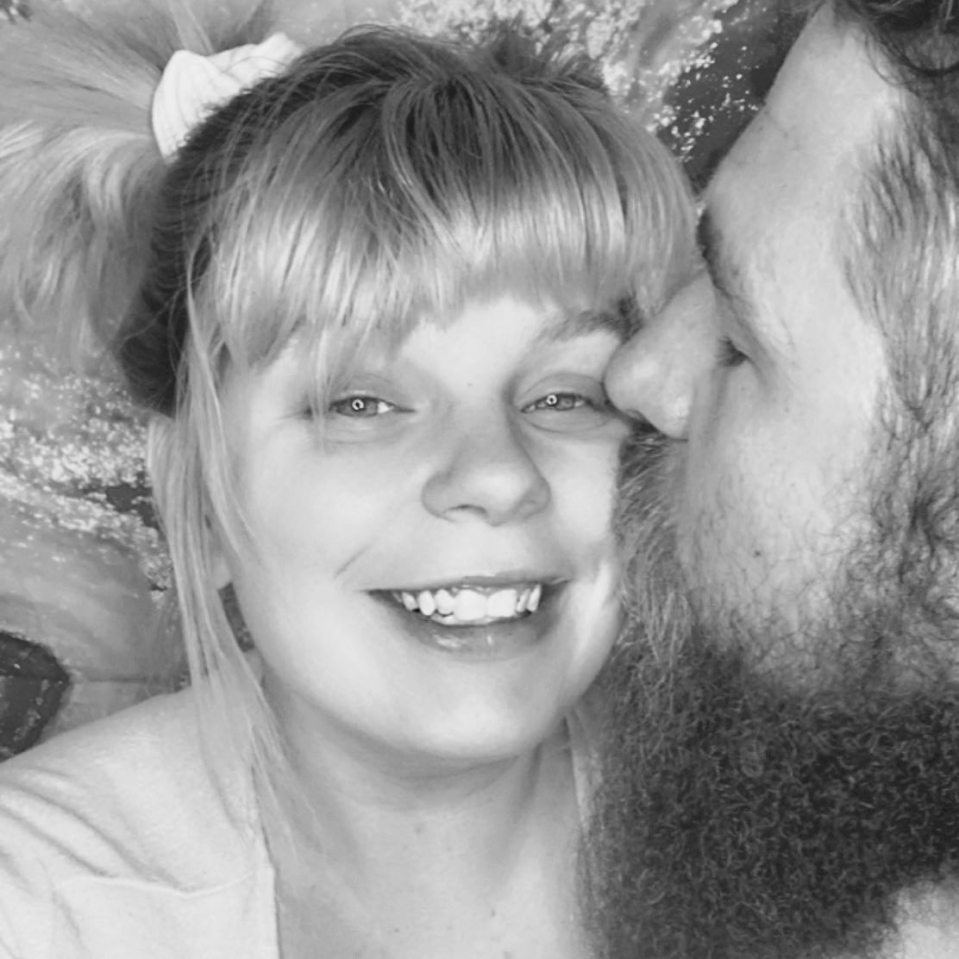 Black and white photo of a couple; a woman with her hair in pigtails smiles as a man kisses her cheek.